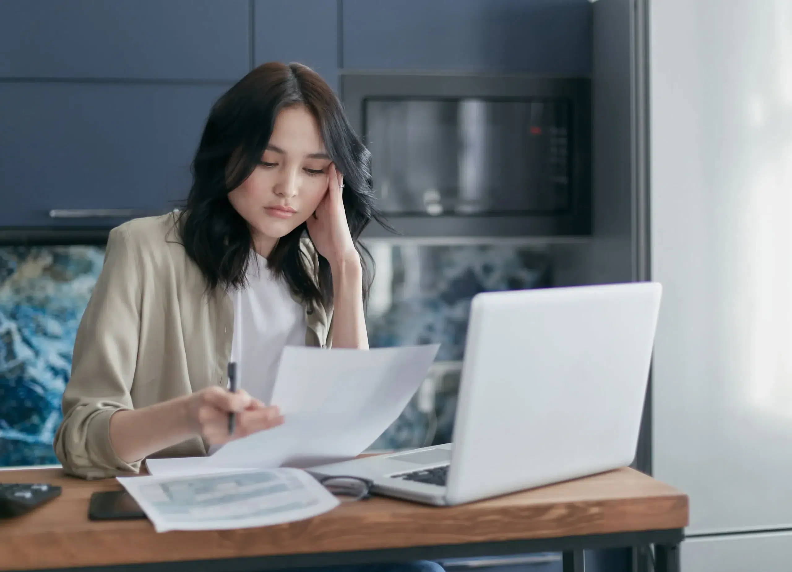 Women holding head while looking at paperwork in front of laptop